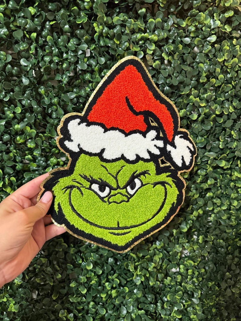 K&ouml;nnte beinhalten: Ein gr&uuml;nes Grinch-Gesicht mit roter Weihnachtsm&uuml;tze und wei&szlig;em Rand. Der Grinch hat einen goldenen Rand und wird vor einem gr&uuml;nen, blattreichen Hintergrund gehalten. Das Gesicht des Grinch hat einen schelmischen Ausdruck.