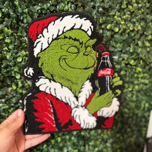 Könnte beinhalten: Ein getufteter Teppich mit einem grünen Charakter, der eine rot-weiße Weihnachtsmannmütze und einen Mantel trägt und eine schwarze Glasflasche mit einem roten Etikett mit der Aufschrift Coca-Cola hält. Das Design hat einen schwarzen Rand.