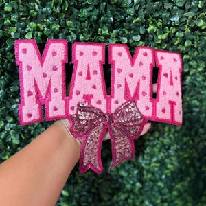 May include: A pink felt sign spelling "MAMA" with heart details. A sequined bow in shades of pink is attached below the letters. The sign is held against a green, leafy backdrop.