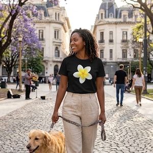 May include: A woman in a black t-shirt with a white and yellow flower design, beige trousers, and a dog lead. A golden retriever is on the lead. The background features a cobbled path, buildings, and trees.