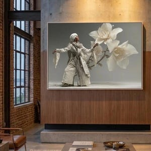 May include: A framed artwork depicting a person in a white outfit and turban, interacting with large white flower blossoms. The artwork is displayed on a wooden wall in a contemporary living room.