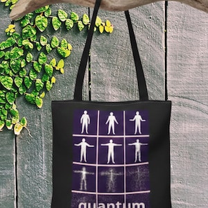 May include: Black tote bag with a graphic of human figures in a grid, with the word "quantum" in white. The bag has black handles and is hanging against a weathered wooden background with green foliage.