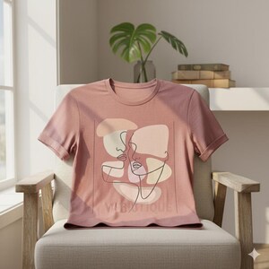 May include: A dusty rose t-shirt featuring a minimalist line art design of two faces. The design includes soft beige and peach-colored shapes with black outlines. The text "VIBUTIQUE" is printed at the bottom of the design.