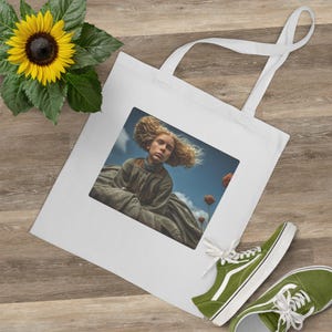 May include: White tote bag featuring a printed image of a person with blonde hair and a vintage dress against a blue sky. The bag has two white handles. A yellow sunflower and green sneakers are also in the image.