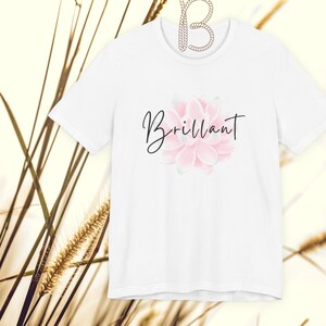 May include: White t-shirt featuring a pink and light green floral design with the word "Brilliant" in a cursive font. The shirt is hanging on a white hanger against a neutral background.
