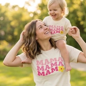 May include: A woman and a child wearing matching cream-colored t-shirts with the word "MAMA" in pink and a yellow flower graphic. The child sits on the woman's shoulders, both smiling in a park setting.