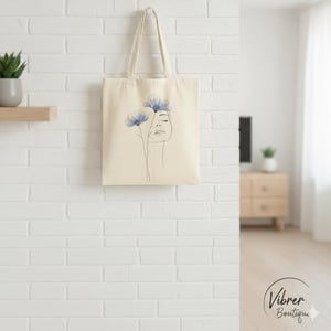 May include: A cream tote bag with a minimalist design. The bag features a line drawing of a woman's face with two blue flowers. The bag has long handles and is hanging against a white brick wall.