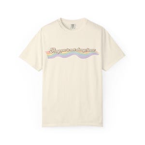 Puede incluir: Camiseta color crema con el texto "Progress is not always linear" en un diseño ondulado de colores arcoíris. La camiseta tiene cuello redondo y mangas cortas. El texto está en una fuente retro.