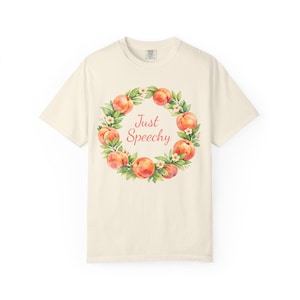 May include: Cream-colored t-shirt featuring a watercolor wreath of peaches and green leaves. The text "Just Speechy" is in the center of the wreath. The shirt has short sleeves and a crew neck.