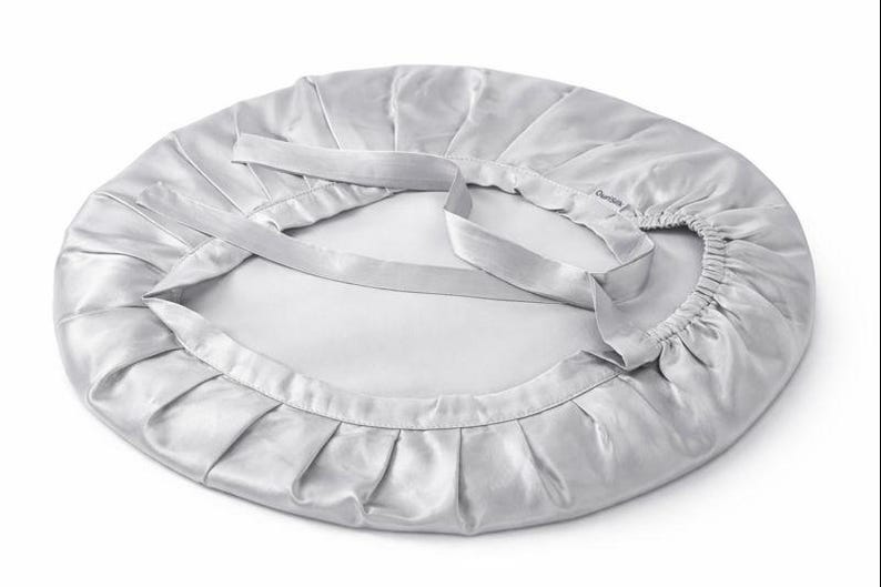 K&ouml;nnte beinhalten: Eine silberne Satinhaube mit ger&uuml;schtem Rand und zwei langen, flachen B&auml;ndern. Die Haube ist kreisf&ouml;rmig und scheint aus einem weichen, gl&auml;nzenden Material zu bestehen. Die B&auml;nder sind zum Binden konzipiert, um die Haube zu sichern.