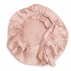 K&ouml;nnte beinhalten: Hellrosa Satin-Haube mit ger&uuml;schtem Rand und Bindeband. Die Haube besteht aus einem gl&auml;nzenden, glatten Stoff und soll das Haar im Schlaf sch&uuml;tzen. Der Markenname "OurSilk" ist sichtbar auf der Haube.