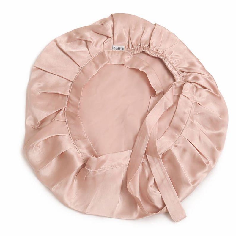 K&ouml;nnte beinhalten: Hellrosa Satin-Haube mit ger&uuml;schtem Rand und Bindeband. Die Haube besteht aus einem gl&auml;nzenden, glatten Stoff und soll das Haar im Schlaf sch&uuml;tzen. Der Markenname "OurSilk" ist sichtbar auf der Haube.