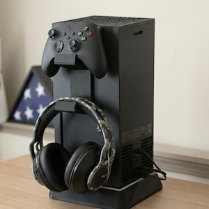 May include: Black gaming console stand with a controller and headset holder. The stand holds a black controller and a pair of black and camouflage headphones. The console is black and has a textured surface.