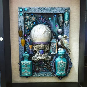 May include: A framed artwork with a central spherical object on an ornate base. Teal and purple bottles, a bird, and other decorative elements surround the sphere. The artwork is enclosed in a black frame with a hinged glass door. The measurements are in centimeters.