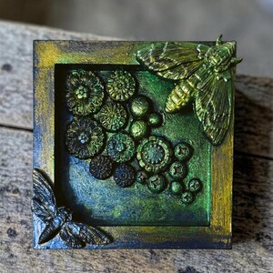 Könnte beinhalten: Ein quadratisches, gerahmtes Kunstwerk mit einem strukturierten, geschichteten Design. Das Kunstwerk zeigt eine Sammlung grüner, kreisförmiger Formen und zwei Mottenfiguren. Der Rahmen hat einen Farbverlauf aus Blau-, Grün- und Goldtönen.