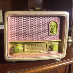 Puede incluir: Una radio de estilo vintage en tonos dorados, rosas y verdes. La radio presenta una cubierta de altavoz de malla, dos perillas verdes y un relieve decorativo de conejo. Las palabras "VOLUME" y "TONE" son visibles.