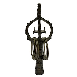 Op de afbeelding: Een donker bronskleurig metalen sculptuur met een centrale verticale paal bekroond door een cirkelvormig frame met een drietandvorm. Vier cirkelvormige metalen ringen hangen aan de dwarsbalk en de basis is voorzien van gegraveerde patronen.