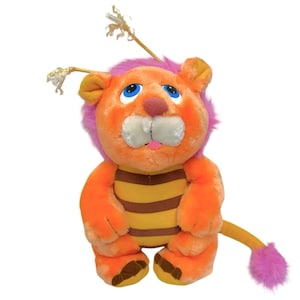 Könnte beinhalten: Ein Plüschtier-Löwe mit orangefarbenem Fell, einer rosa Mähne und einem gestreiften gelb-braunen Bauch. Der Löwe hat blaue Augen, eine rosa Zunge und einen Schwanz mit einem rosa Bommel. Er hat goldene Antennen.