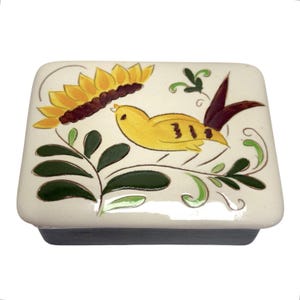 May include: A rectangular ceramic box with a cream-colored lid featuring a hand-painted design of a yellow bird, a large yellow sunflower with a dark brown center, and dark green leaves with thin brown outlines on a smooth, glossy surface.