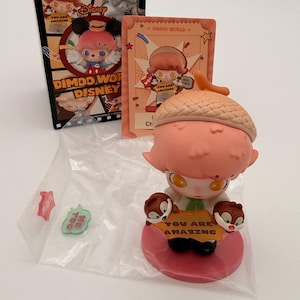 May include: A DIMOO WORLD Disney blind box figure. The figure has a pink head and a brown acorn hat. It holds a sign that says "YOU ARE AMAZING". The box and card feature Disney characters.
