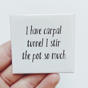 May include: A square white magnet featuring black text that reads, I have carpal tunnel I stir the pot so much. The magnet has a smooth, glossy finish and clean, rounded corners, presenting a simple design with bold, legible lettering.