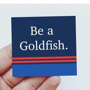 May include: A square sticker with a dark blue background featuring the white text Be a Goldfish. Below the text are two horizontal red stripes, followed by a solid light blue band at the bottom edge of the square sticker design.