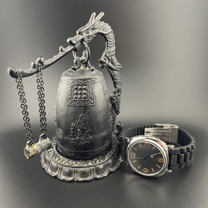 May include: A silver-colored decorative bell with a dragon-shaped handle and a black rubber watch with a silver case and black dial. The bell has intricate carvings and hangs from a chain.