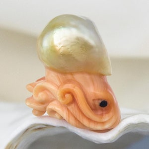 May include: A carved coral octopus with a large, iridescent pearl on top. The octopus is a light orange colour with a black eye. The pearl is a pale yellow with a green iridescent sheen.