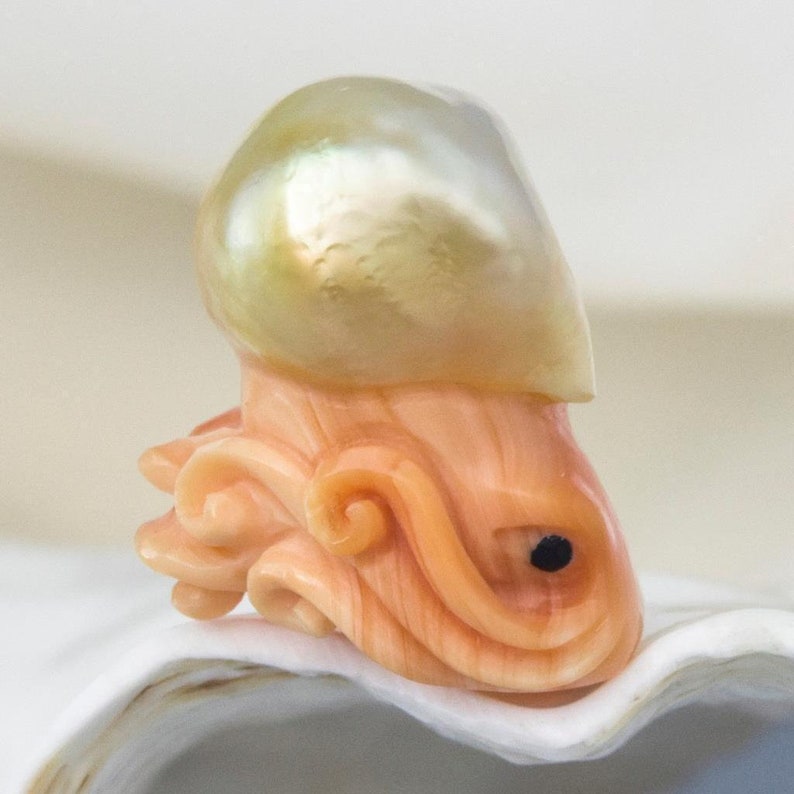 May include: A carved coral octopus with a large, iridescent pearl on top. The octopus is a light orange colour with a black eye. The pearl is a pale yellow with a green iridescent sheen.