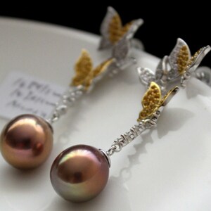 Puede incluir: Un par de pendientes de plata con una gota de perla marr&oacute;n y un dije de mariposa con detalles dorados y transparentes.