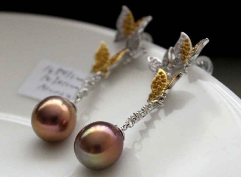 Puede incluir: Un par de pendientes de plata con una gota de perla marr&oacute;n y un dije de mariposa con detalles dorados y transparentes.