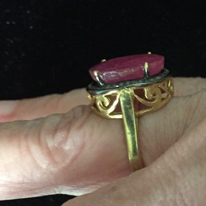 May include: A gold ring with a large, oval-shaped ruby gemstone set in a filigree design. The ruby is a deep pink color and is surrounded by smaller diamonds.
