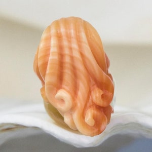 May include: A carved coral sculpture of a woman's head with long, flowing hair. The coral is a light orange colour with white and pink streaks.