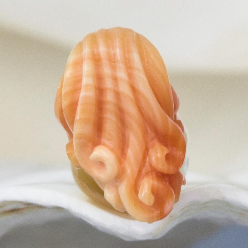 May include: A carved coral sculpture of a woman's head with long, flowing hair. The coral is a light orange colour with white and pink streaks.