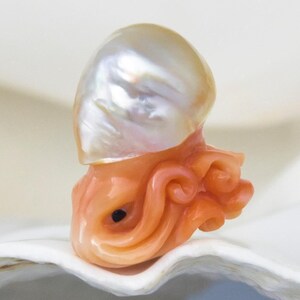 May include: A white baroque pearl with a pink coral octopus carving. The pearl is on top of the octopus and the octopus is sitting on a white shell.