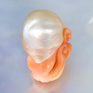 May include: A large, white, pearlescent, irregularly shaped pearl set in a carved, orange coral base.