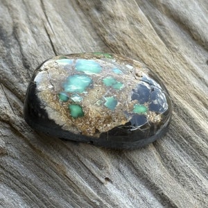 May include: A green and brown cabochon gemstone with a black backing. The stone has a mottled pattern with green and brown spots.
