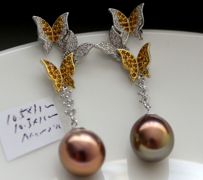 Puede incluir: Un par de pendientes de plata con una perla colgante y tres abalorios de mariposa. Las mariposas son doradas con piedras amarillas y transparentes.