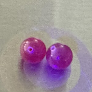 May include: Two round, pink ruby beads with a slight purple hue under UV light.
