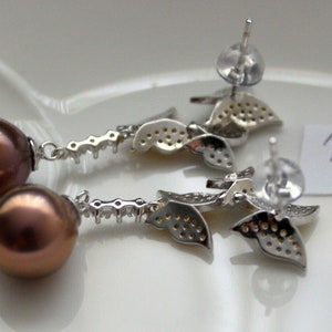 Puede incluir: Un par de pendientes de plata con una perla marr&oacute;n y un dise&ntilde;o de hojas. Las hojas est&aacute;n incrustadas con peque&ntilde;as piedras transparentes.
