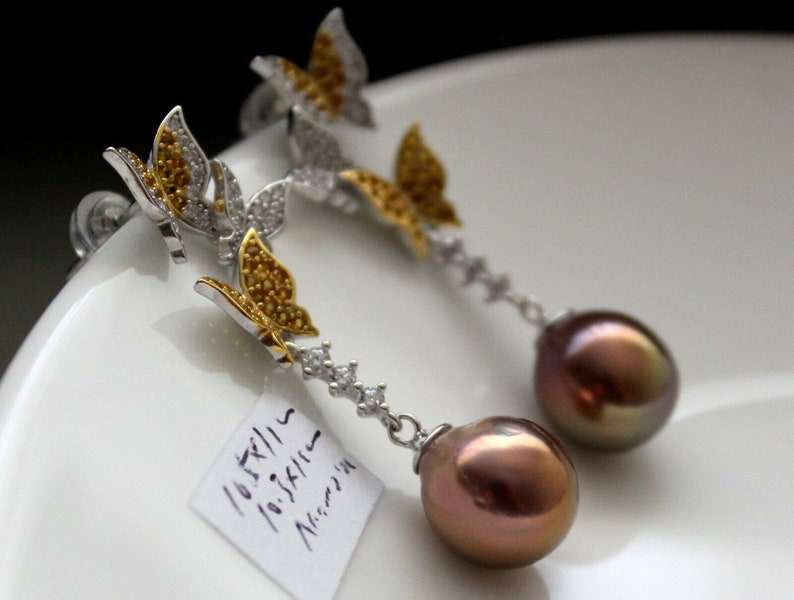 Puede incluir: Un par de pendientes de plata con un dise&ntilde;o de mariposa y una perla marr&oacute;n. Las mariposas est&aacute;n adornadas con piedras amarillas y transparentes.