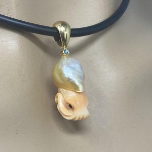 May include: A gold pendant featuring a carved octopus with a large, golden pearl on top. The pearl has a white and gold iridescent sheen. The pendant is hanging from a black cord.