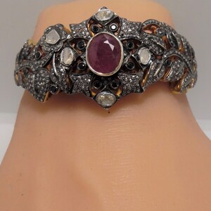 May include: A silver and gold bracelet with a large red ruby gemstone surrounded by smaller white and black diamonds. The bracelet has a floral design.