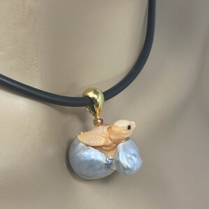 K&ouml;nnte beinhalten: Eine goldene Halskette mit einem Anh&auml;nger, der eine geschnitzte Schildkr&ouml;te zeigt, die aus einer Perlmuschel herauskommt. Die Schildkr&ouml;te ist aus einem hellorangen Material gefertigt und hat einen rosafarbenen Edelstein auf dem R&uuml;cken. Die Halskette ist an einem schwarzen Band befestigt.