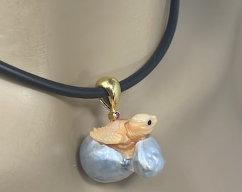 May include: A gold pendant necklace featuring a carved turtle emerging from a pearl shell. The turtle is crafted from a light orange material and has a pink gemstone on its back. The necklace is on a black cord.