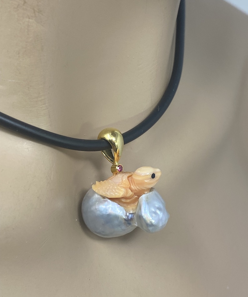 K&ouml;nnte beinhalten: Eine goldene Halskette mit einem Anh&auml;nger, der eine geschnitzte Schildkr&ouml;te zeigt, die aus einer Perlmuschel herauskommt. Die Schildkr&ouml;te ist aus einem hellorangen Material gefertigt und hat einen rosafarbenen Edelstein auf dem R&uuml;cken. Die Halskette ist an einem schwarzen Band befestigt.