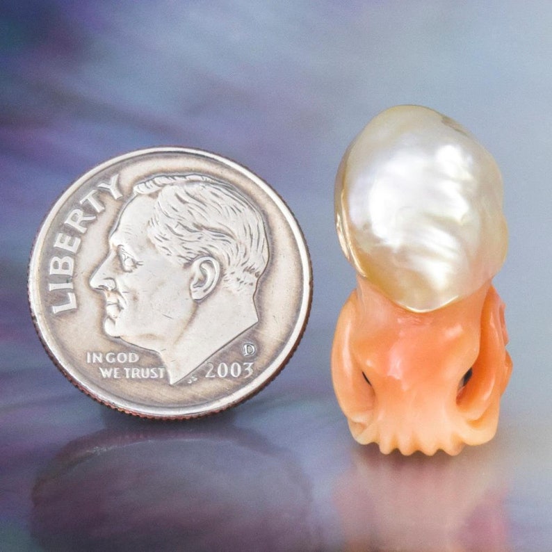 May include: A white baroque pearl with a golden hue, carved into the shape of an octopus, with coral tentacles. The pearl is about the size of a UK ten pence coin.