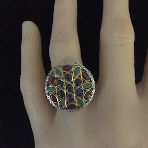 May include: A gold ring with a round design featuring a cluster of oval-shaped gemstones in shades of blue, green, and red. The ring is set with smaller white gemstones around the perimeter.