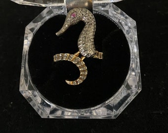 May include: A gold ring with a detailed, diamond-encrusted seahorse design. The seahorse has a red gemstone for an eye.