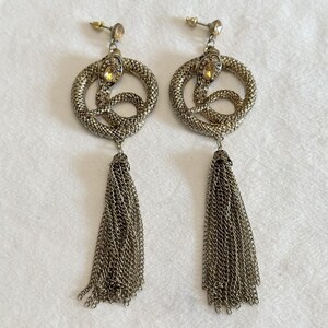May include: A pair of silver-toned dangle earrings. Each earring features a coiled snake design with a yellow gemstone and a chain tassel. The earrings are designed to be worn with a post and backing.